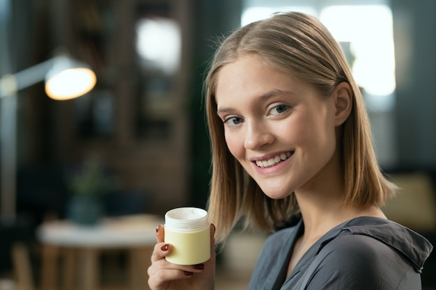 Giovane bella donna con i capelli biondi che tiene in mano un vasetto con un prodotto cosmetico naturale fatto a mano e ti guarda con un sorriso a trentadue denti