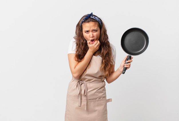 Giovane bella donna con la bocca e gli occhi spalancati e la mano sul concetto di chef sul mento e con in mano una padella