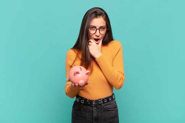 Giovane bella donna con la bocca e gli occhi spalancati e la mano sul mento, sentendosi spiacevolmente scioccata, dicendo cosa o wow. concetto di salvadanaio