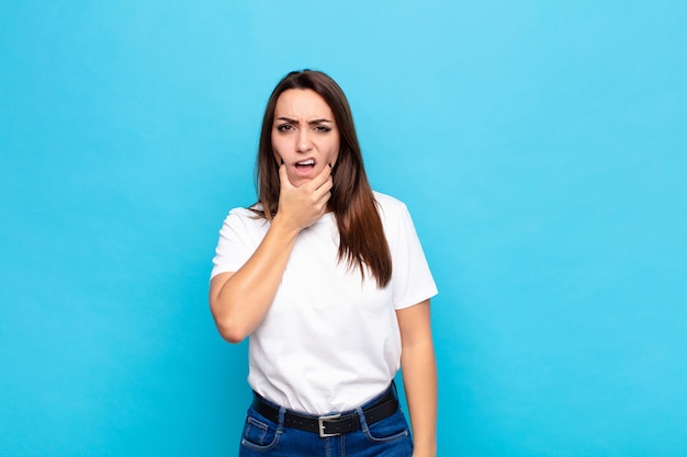 Giovane bella donna con la bocca e gli occhi spalancati e la mano sul mento, sentendosi spiacevolmente scioccata, dicendo cosa o wow contro il muro blu