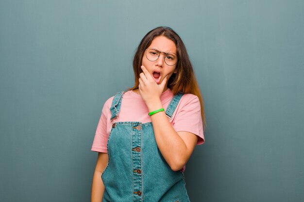 Giovane bella donna con la bocca e gli occhi spalancati e la mano sul mento, sentendosi spiacevolmente scioccata, dicendo cosa o wow contro il muro del grunge