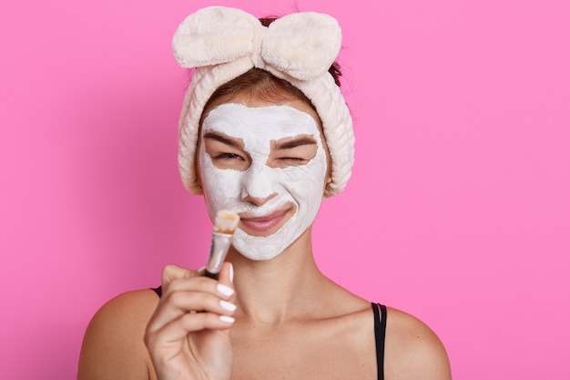 Giovane bella donna con la maschera facciale di argilla sul viso tenendo la spazzola nelle mani dopo aver fatto il trattamento di bellezza, facendo una faccia buffa mentre fa la cura della pelle, divertirsi a casa.