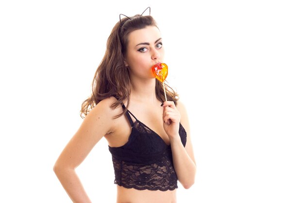 Giovane bella donna con le orecchie di gatto sulla testa che indossa un reggiseno di pizzo nero che mangia caramelle a forma di cuore