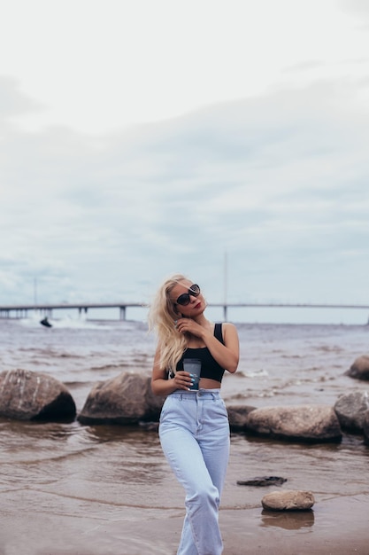 Giovane bella donna con lunghi capelli biondi che guarda il mare e beve caffè