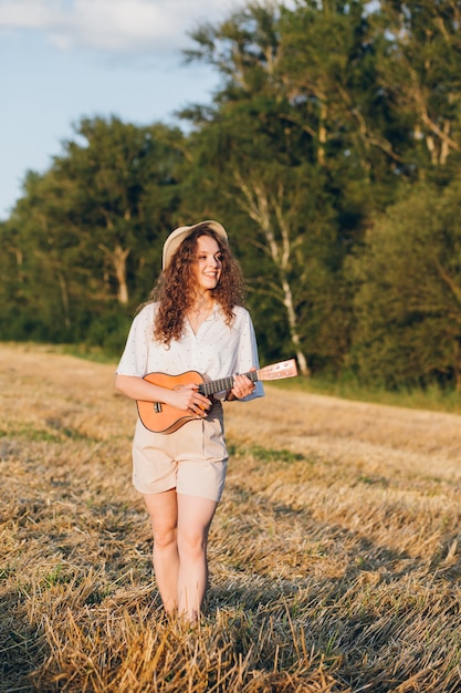 Giovane bella donna con lunghi capelli ricci in pantaloncini corti, un cappello con una chitarra si pone in un campo di grano in estate al tramonto