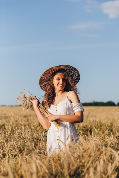 Giovane bella donna con lunghi capelli ricci pone in un campo di grano in estate al tramonto