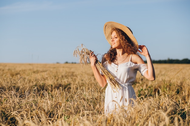 Giovane bella donna con lunghi capelli ricci pone in un campo di grano in estate al tramonto