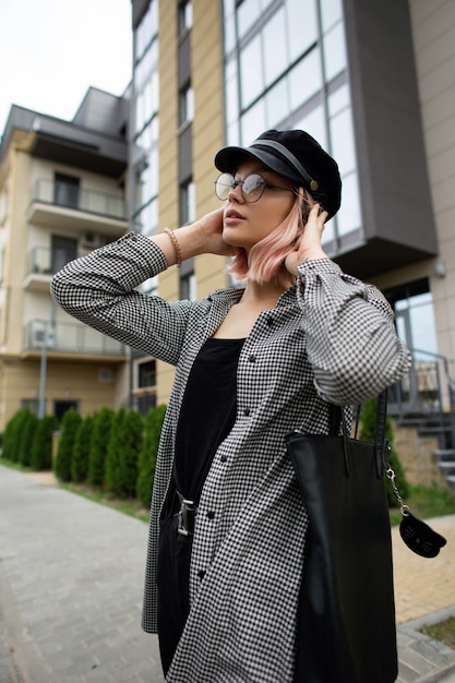 Giovane bella donna con occhiali vintage e un cappello in una camicia a quadri alla moda con una borsa in pelle nera cammina per strada vicino all'edificio