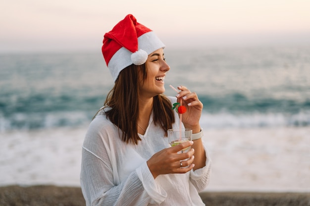 Giovane bella donna con un cappello da Babbo Natale con un bicchiere di cocktail in mano cammina sulla spiaggia