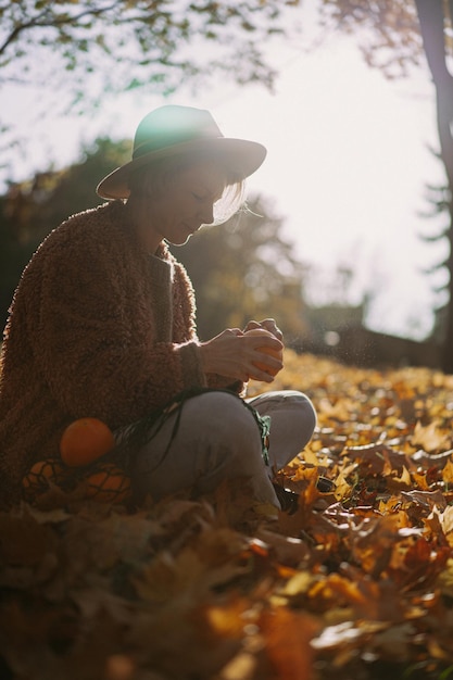 Giovane bella donna con un cappello in un parco autunnale, una borsa di corda con arance, una donna vomita foglie d'autunno. Umore autunnale, colori vivaci della natura.