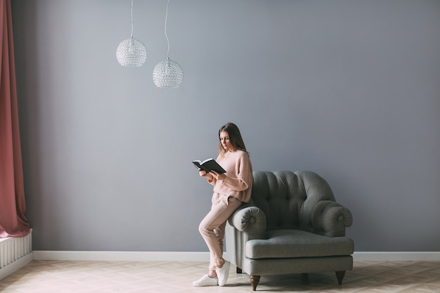 Giovane bella donna con un libro vicino alla sedia in casa.