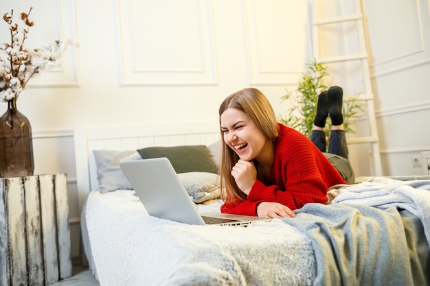 Giovane bella donna con un maglione rosso che scrive su un computer portatile seduto su un letto in una stanza luminosa. Lavorare da remoto in un'atmosfera casalinga durante la quarantena