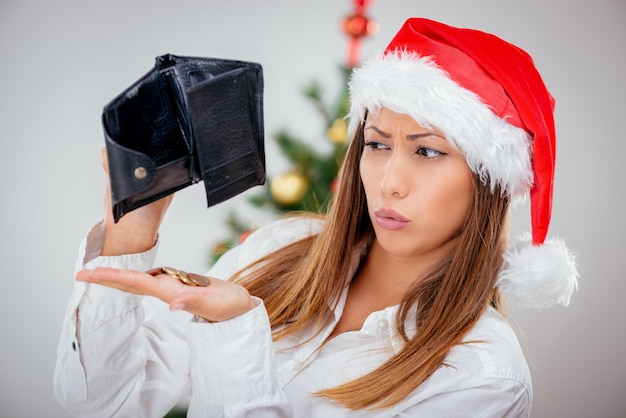 Giovane bella donna d'affari che indossa il cappello di Babbo Natale e guarda preoccupato per il portafoglio vuoto.