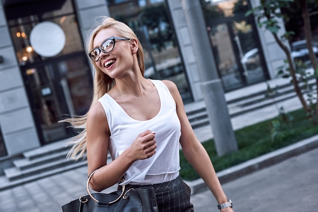 Giovane bella donna d'affari con gli occhiali da sole che sorride e cammina vicino all'edificio degli affari che lei è a