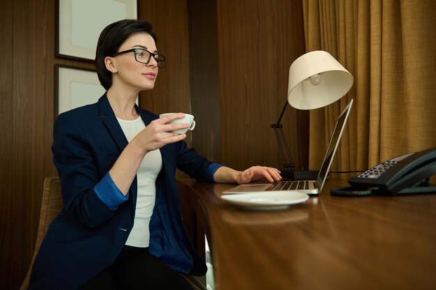 Giovane bella donna d'affari, imprenditrice, responsabile delle vendite di successo in occhiali e abito casual da lavoro si siede alla scrivania e tiene una tazza di caffè mentre lavora al computer portatile nella camera d'albergo durante il viaggio d'affari