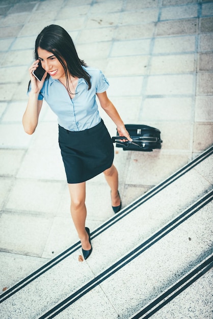 Giovane bella donna d'affari in piedi all'aperto con la valigia pronta per un viaggio d'affari e parlando al cellulare.