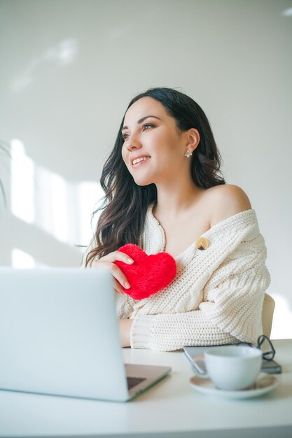 Giovane bella donna dai capelli scuri di 34 anni in maglione lavorato a maglia sta parlando al videofonino e tiene in mano un cuore di peluche San Valentino La modella ha ricevuto un regalo