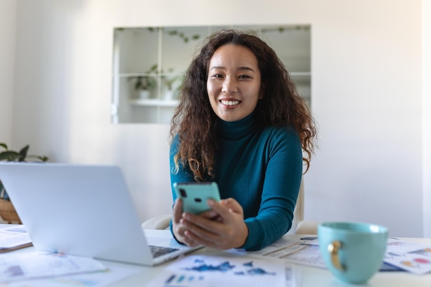 Giovane bella donna di affari asiatica nell'ufficio che si siede nella sedia dell'ufficio che parla sul suo smartphone