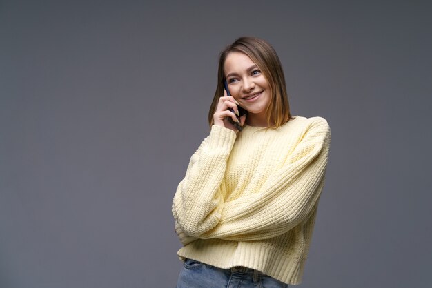 Giovane bella donna di etnia caucasica che parla al telefono con un maglione volumetrico giallo su una superficie grigia con occhi azzurri