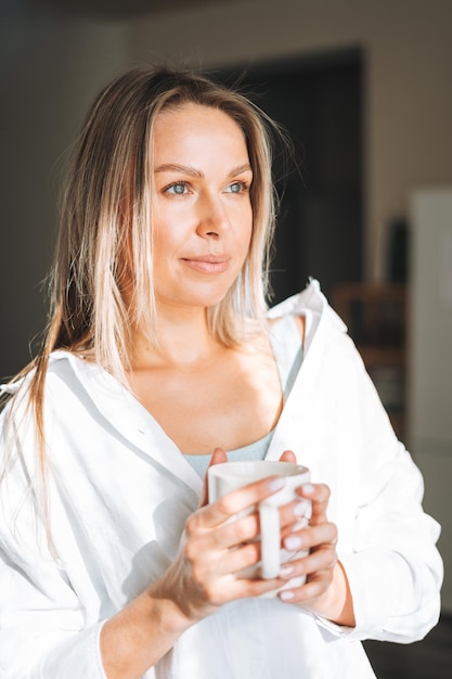 Giovane bella donna di quarant'anni con lunghi capelli ricci biondi in camicia bianca con una tazza di tè in mano vicino alla finestra con una luce intensa in interni luminosi a casa