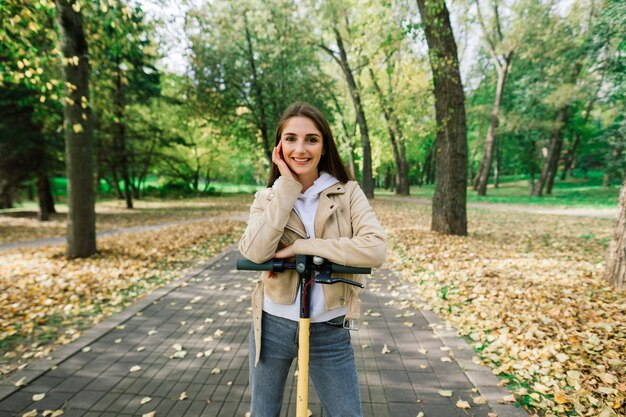 Giovane bella donna elegante in sella a uno scooter elettrico dal parco autunnale, trasporto ecologico