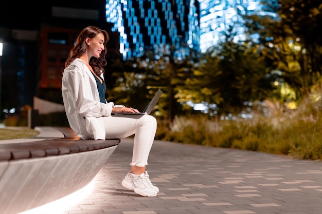 Giovane bella donna europea che utilizza il computer portatile nel parco cittadino di notte seduto sul lato della panchina