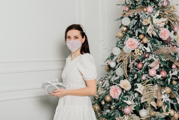 Giovane bella donna europea in maschera medica con regali di festa a casa, coronavirus in quarantena.