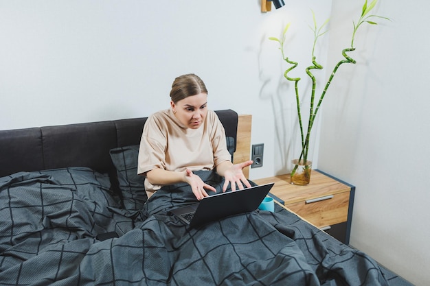 Giovane bella donna europea seduta sul letto con il caffè che lavora con il computer a casa lavoro remoto Donna felice e sorridente a letto a casa