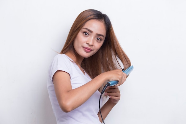 Giovane bella donna felice asiatica che raddrizza i capelli con la piastra per capelli su fondo bianco