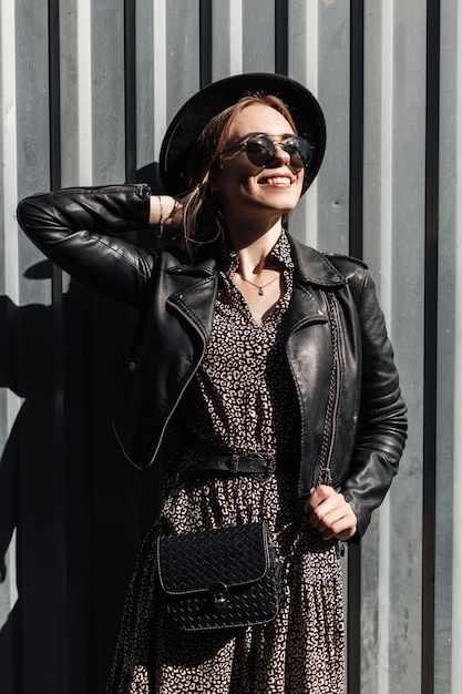 Giovane bella donna felice con occhiali neri in abiti alla moda con abito vintage e giacca di pelle con borsa vicino a sfondo metallico sulla strada