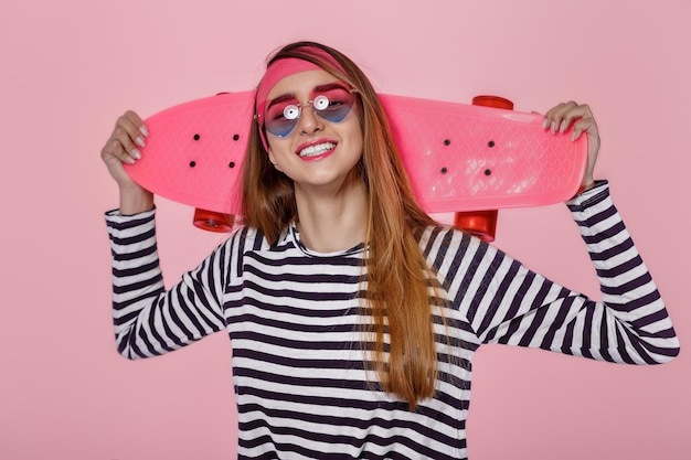 Giovane bella donna felice in bicchieri con skateboard su sfondo rosa