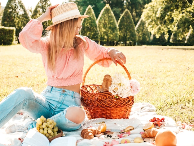 Giovane bella donna hipster in jeans estivi alla moda, maglietta rosa e cappello. Donna spensierata che fa picnic all'esterno.