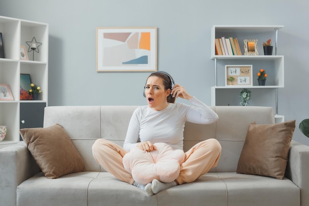 Giovane bella donna in abbigliamento casual con le cuffie seduto su un divano a casa interno guardando la telecamera essendo confuso cercando di sentire qualcosa che si toglie le cuffie