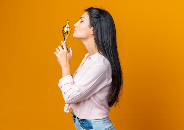 Giovane bella donna in abiti casual che tiene in mano un trofeo che lo bacia felice e fiducioso in piedi lateralmente sull'arancia