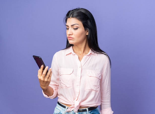 Giovane bella donna in abiti casual che tiene smartphone guardandolo con una faccia seria