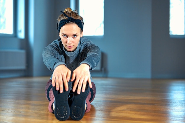 Giovane bella donna in abiti sportivi che fa allungamento mentre sedendosi sul pavimento davanti alla finestra in palestra