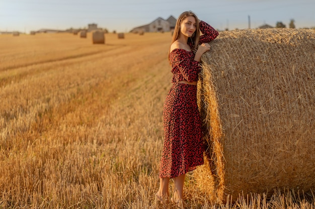 Giovane bella donna in abito rosso da vicino sullo sfondo del campo con mucchi di fieno all'ora del tramonto.