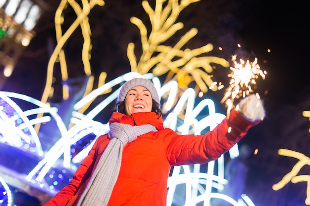 Giovane bella donna in berretto lavorato a maglia e sciarpa in piedi in città con luce bengala, sparkler. Celebrazione di concetto e natale.