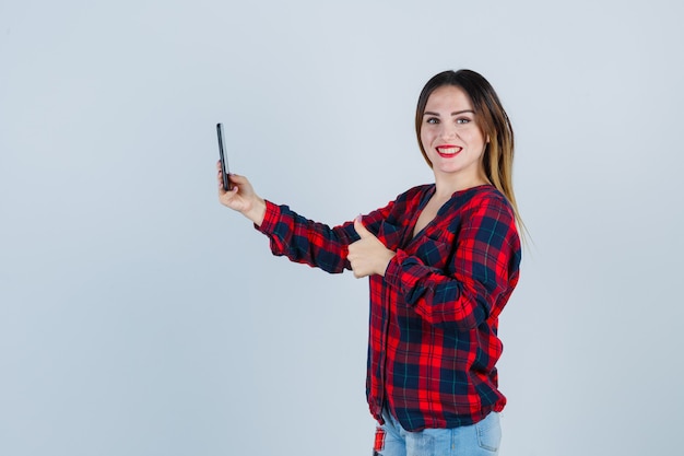 Giovane bella donna in camicia casual prendendo selfie, mostrando pollice in su e guardando beata, vista frontale.