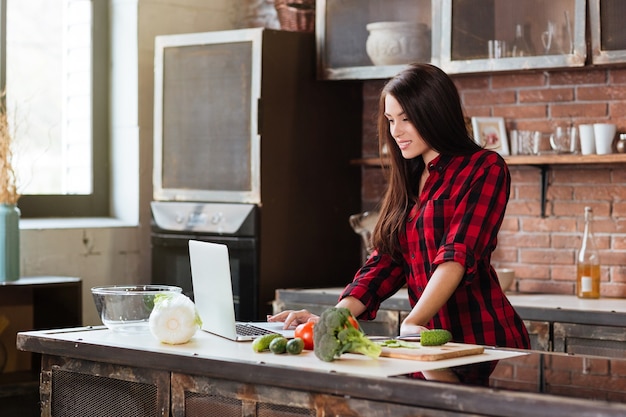 Giovane bella donna in camicia rossa in piedi al tavolo in cucina con il computer portatile e alla ricerca di ricette.