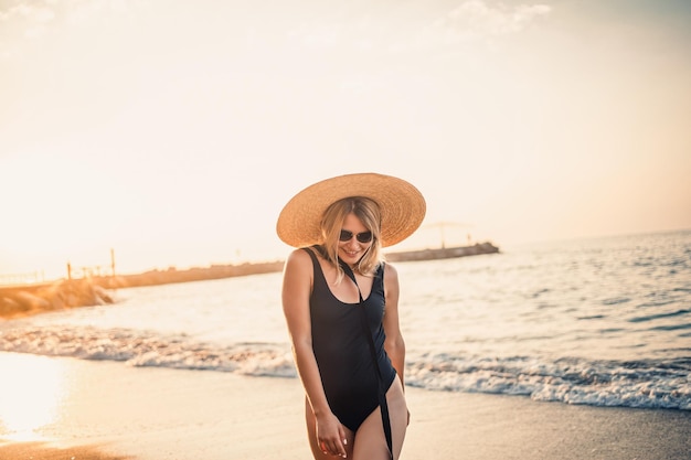 Giovane bella donna in costume da bagno nero e cappello con occhiali cammina lungo la spiaggia in Turchia al tramonto Il concetto di ricreazione in mare Messa a fuoco selettiva