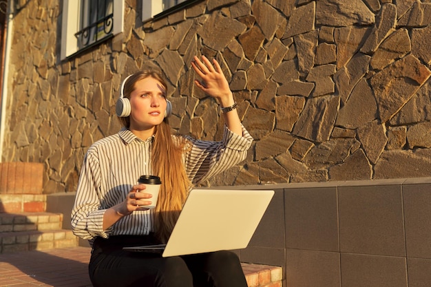 Giovane bella donna in cuffia con laptop che riposa seduto sui gradini in città, ascolta musica guardando video, coprendo il viso dal sole con la mano. Felicità, relax, stile di vita, tecnologia giovanile