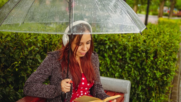 Giovane bella donna in cuffie con ombrello trasparente che legge un libro seduto su una panchina nel parco cittadino sotto la pioggia Rilassata donna appartata che si rilassa con la musica del libro che si ripara con l'ombrello dalla pioggia