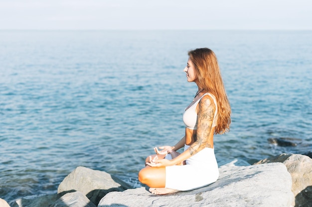 Giovane bella donna in forma con i capelli lunghi in abiti sportivi bianchi pratica yoga e godersi la vita sulla spiaggia del mare salute mentale e fisica