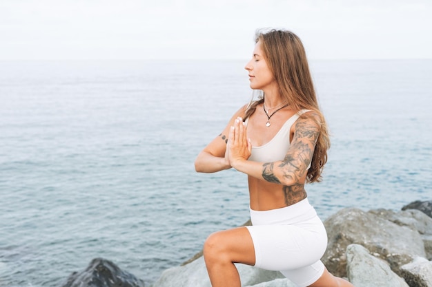 Giovane bella donna in forma con i capelli lunghi in abiti sportivi bianchi pratica yoga e meditazione sul mare