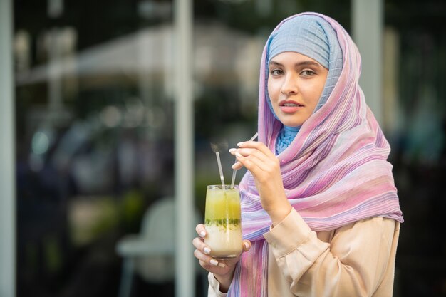 Giovane bella donna in hijab che beve cocktail di raffreddamento dal vetro mentre si riposa all'aperto