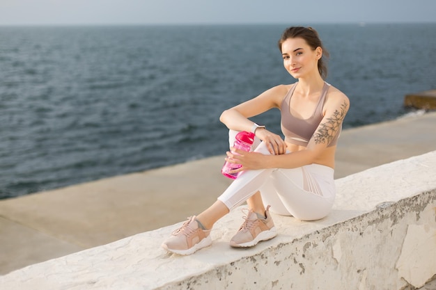 Giovane bella donna in top sportivo e leggings bianchi con in mano una bottiglia sportiva rosa mentre guarda felicemente in camera con vista mare sullo sfondo