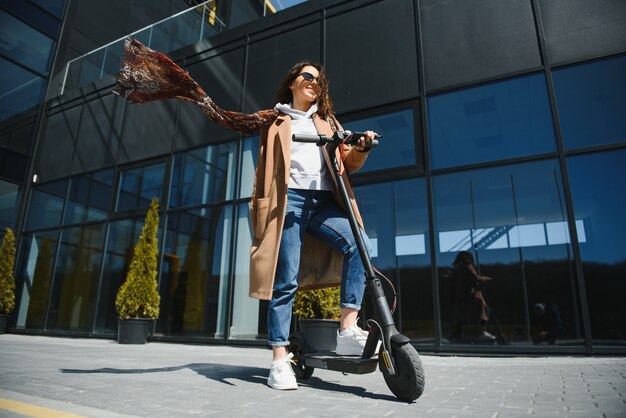 Giovane bella donna in una giacca sorride e cavalca uno scooter elettrico per lavorare