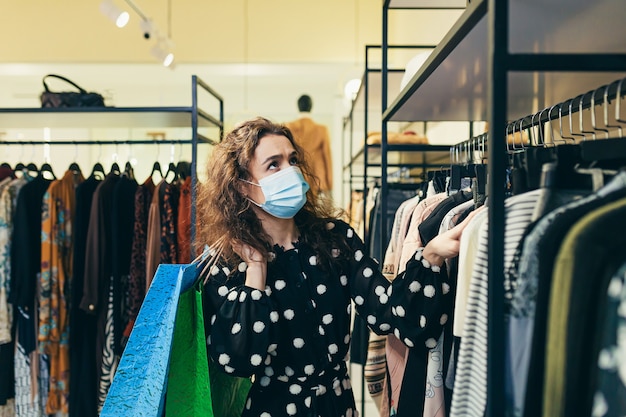 Giovane bella donna in una maschera protettiva sul viso, sceglie i vestiti nel negozio