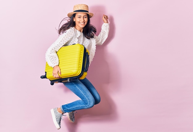 Giovane bella donna in vacanza indossando abiti estivi e cappello sorridendo felice Saltando con il sorriso sul viso tenendo la borsa da cabina su sfondo isolato